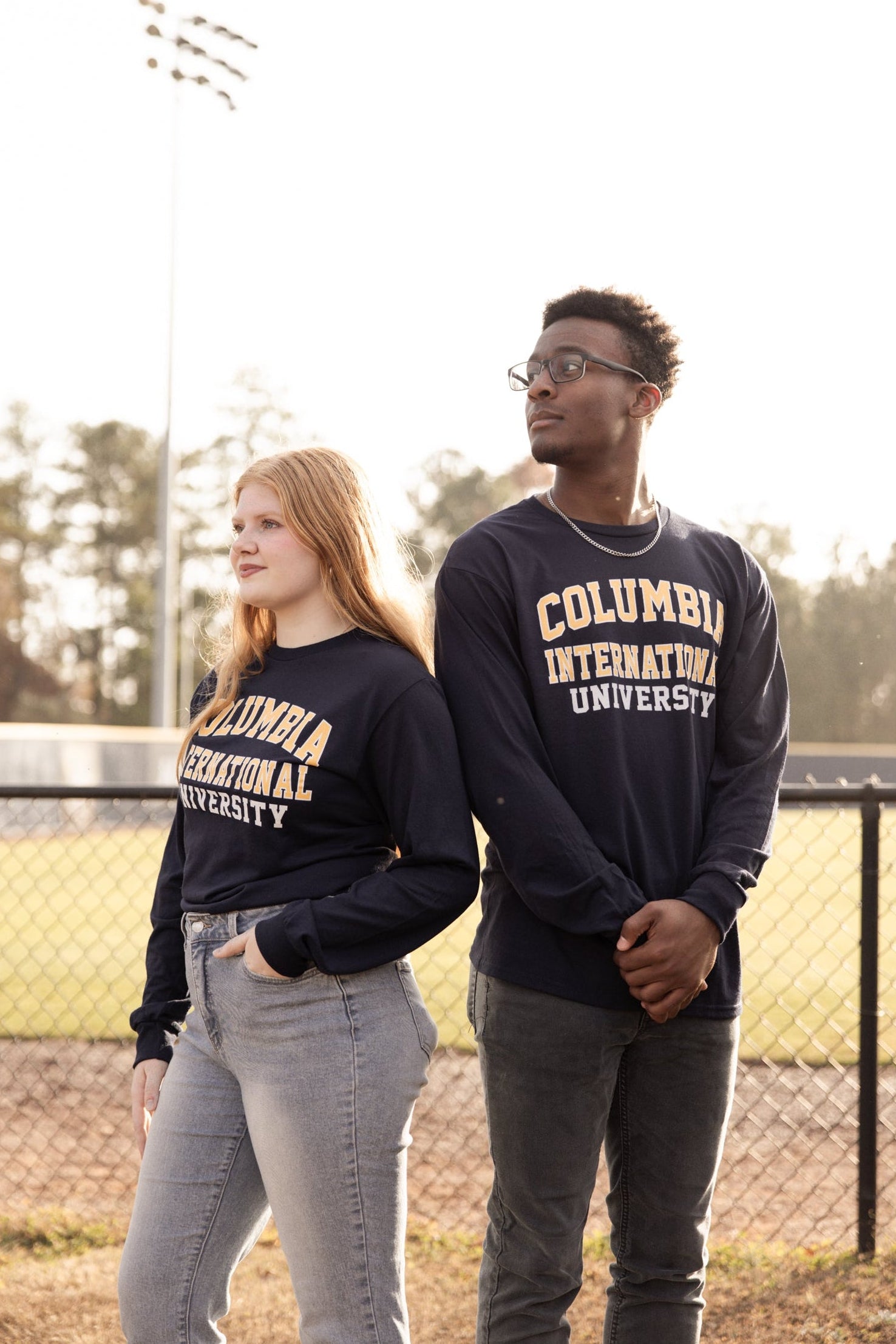 Two people wearing Columbia International University long-sleeve shirts outdoors near a baseball field.