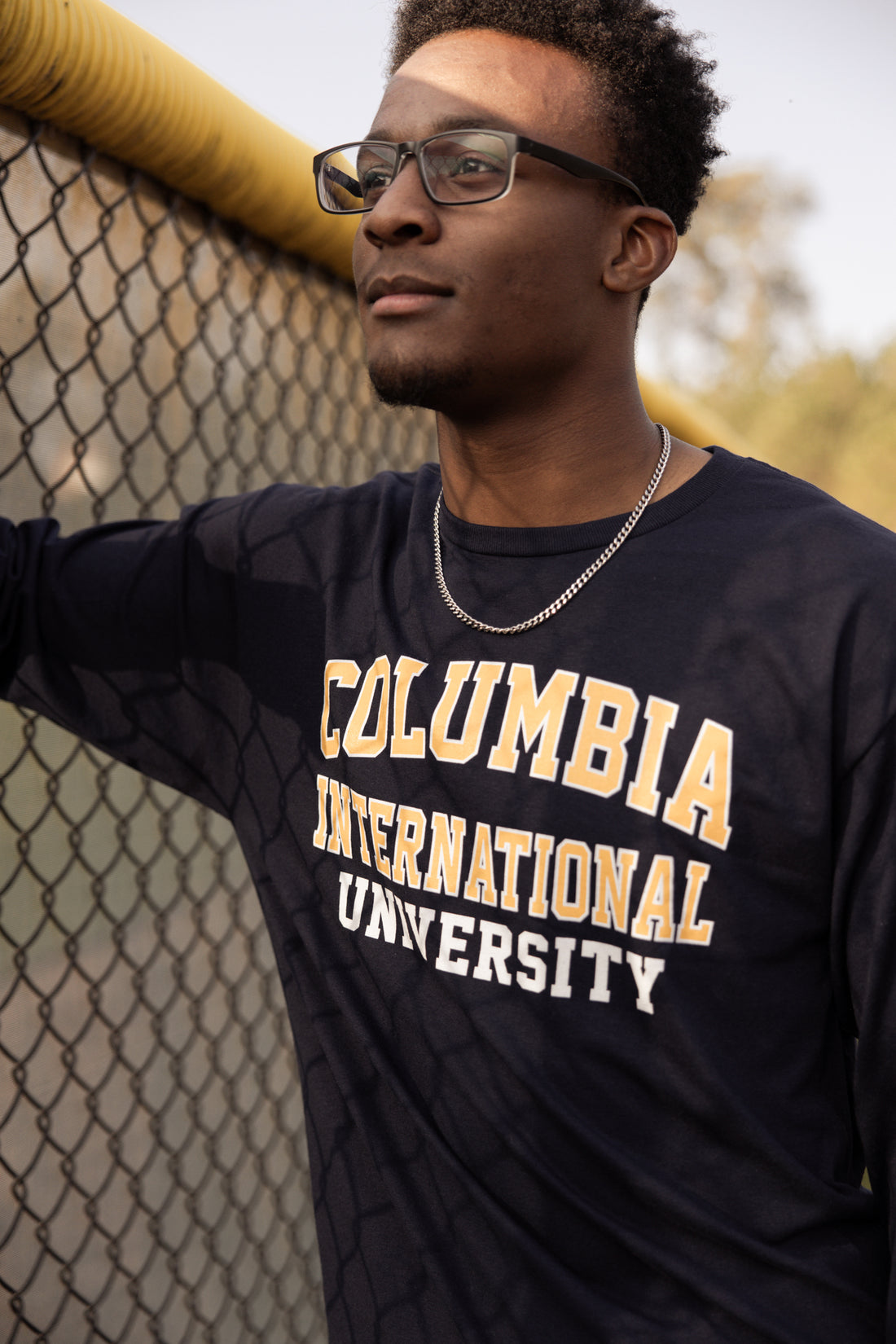 Close-up of a man wearing a Columbia International University long-sleeve shirt leaning against a chain-link fence at a baseball field.