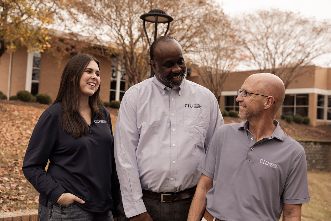 Three people smiling and standing outdoors in CIU professional merch with trees and campus buildings in the background.