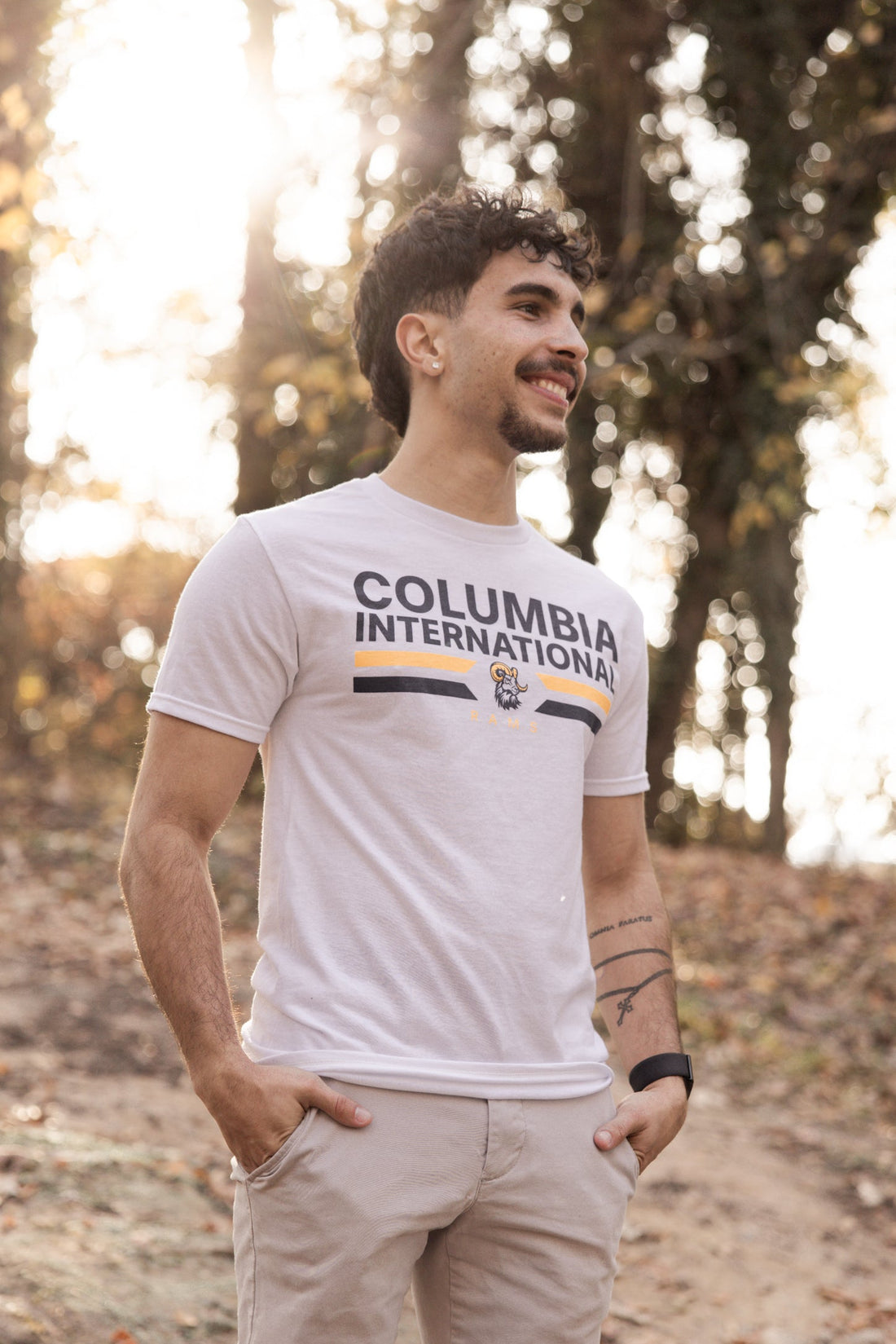 Man wearing a white Columbia International Rams shirt standing with trees in the background.