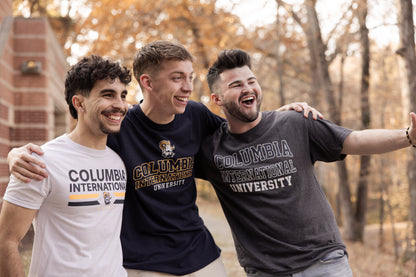 Three men wearing Columbia University shirts smiling, with trees in the background.