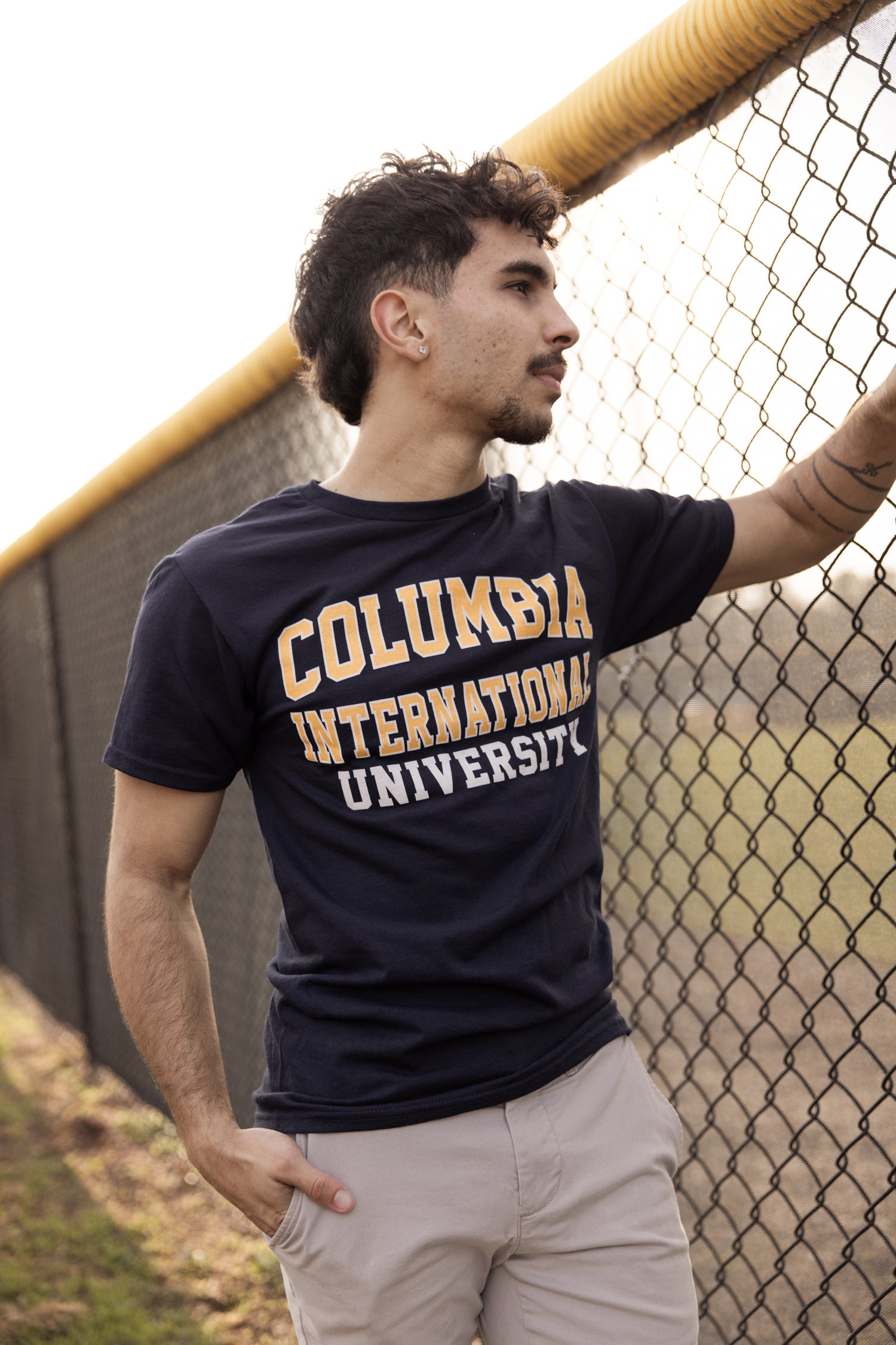 Man wearing a navy Columbia International University t-shirt leaning against a fence.