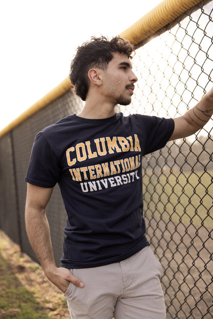 Man wearing a navy Columbia International University t-shirt leaning against a fence.