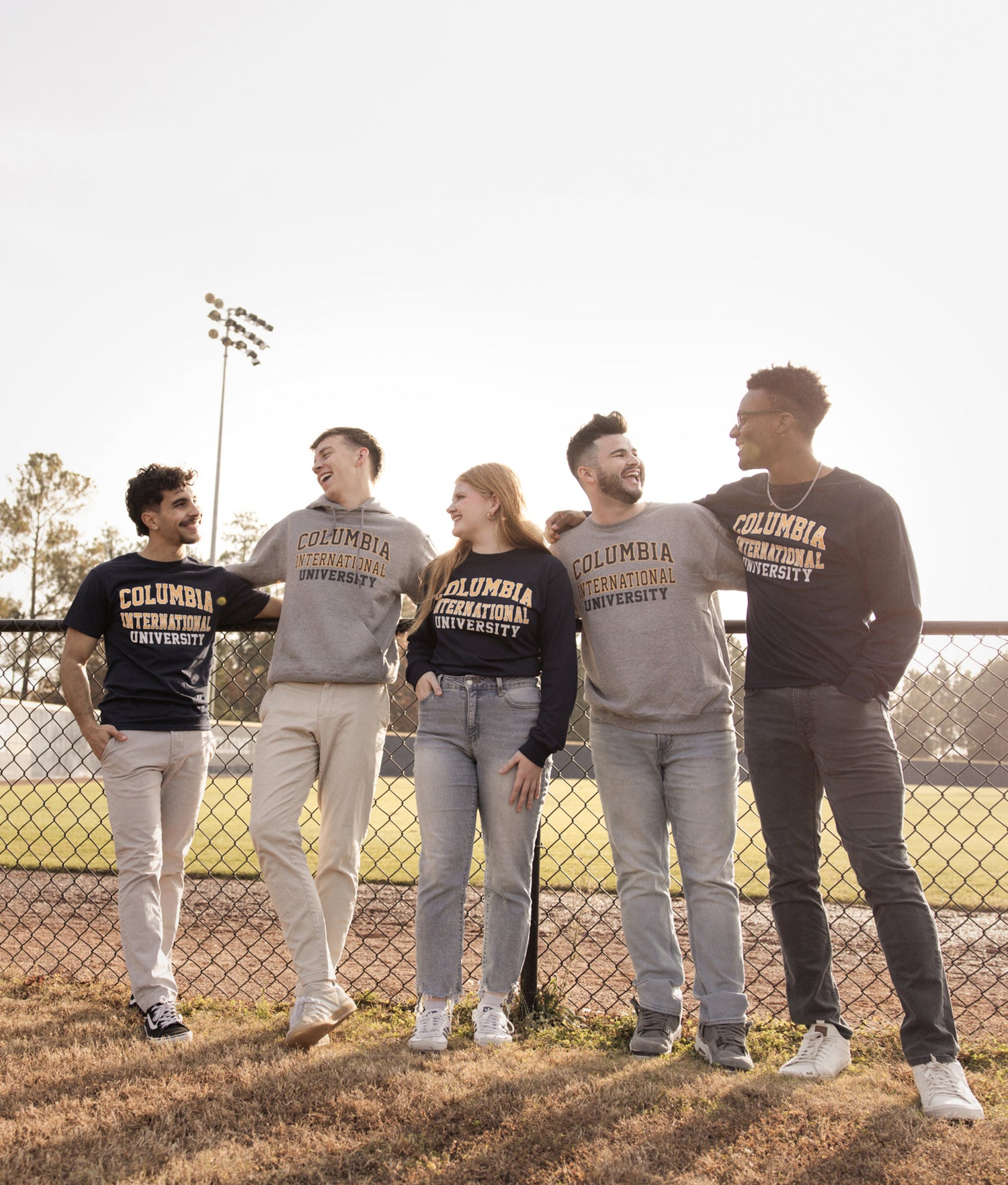 Group of five people wearing Columbia International University merch standing outdoors near a baseball field.