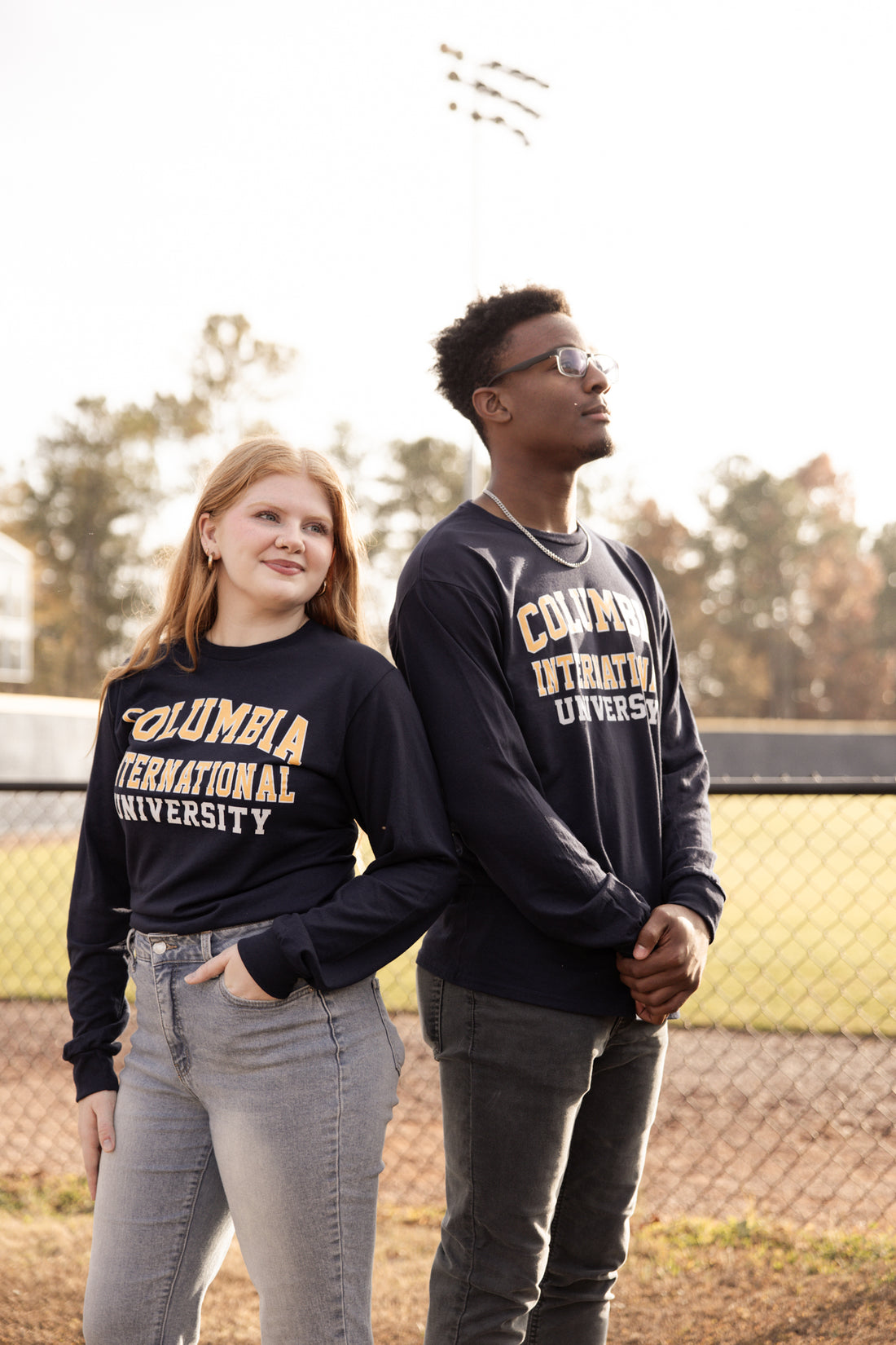 Two people wearing Columbia International University long-sleeve shirts outdoors near a baseball field.