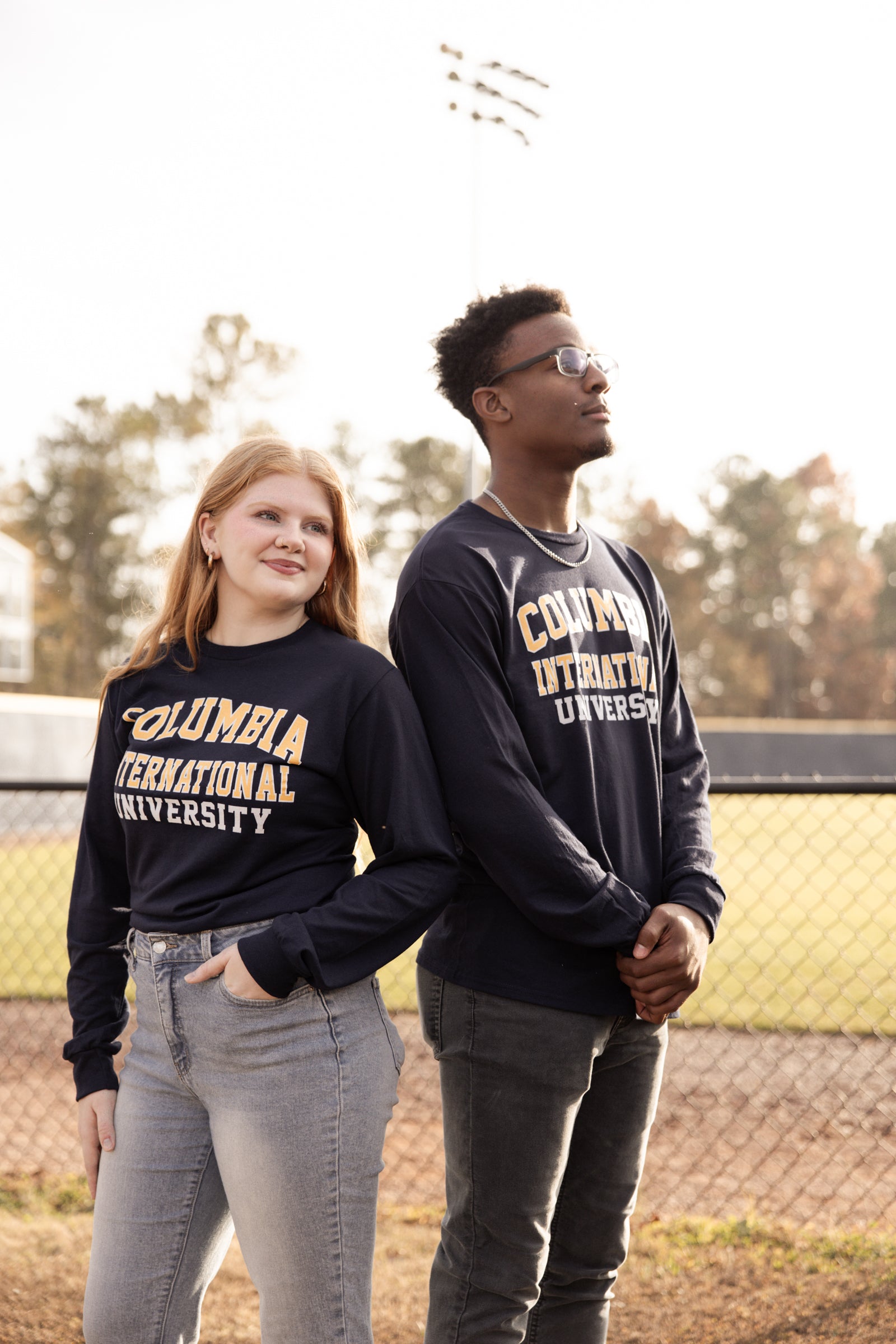 Two people wearing Columbia International University long-sleeve shirts outdoors near a baseball field.